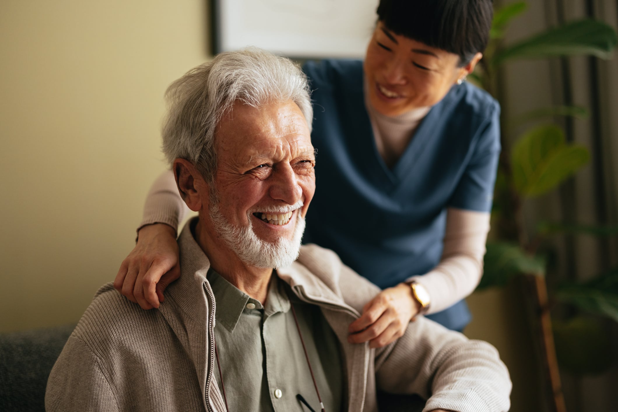 Close up shot of a senior man sitting and looking ahead of him, smiling. There is a female carer helping him take off his jacket.