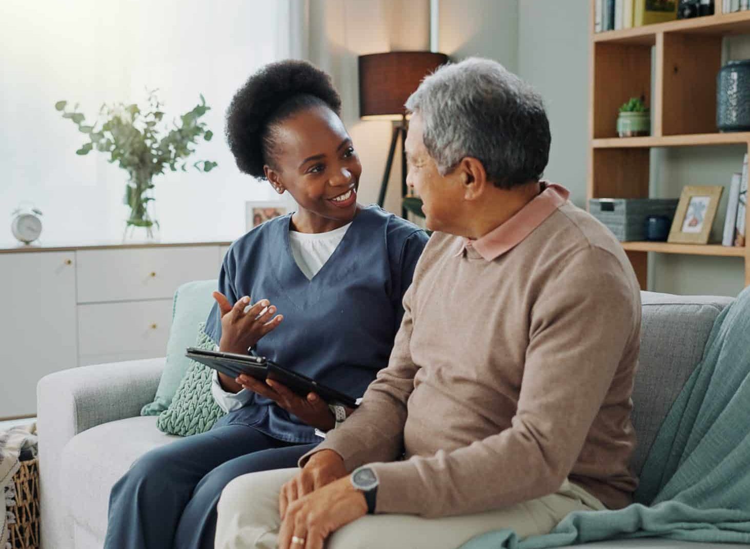 Woman wearing scrubs sitting by senior man in home